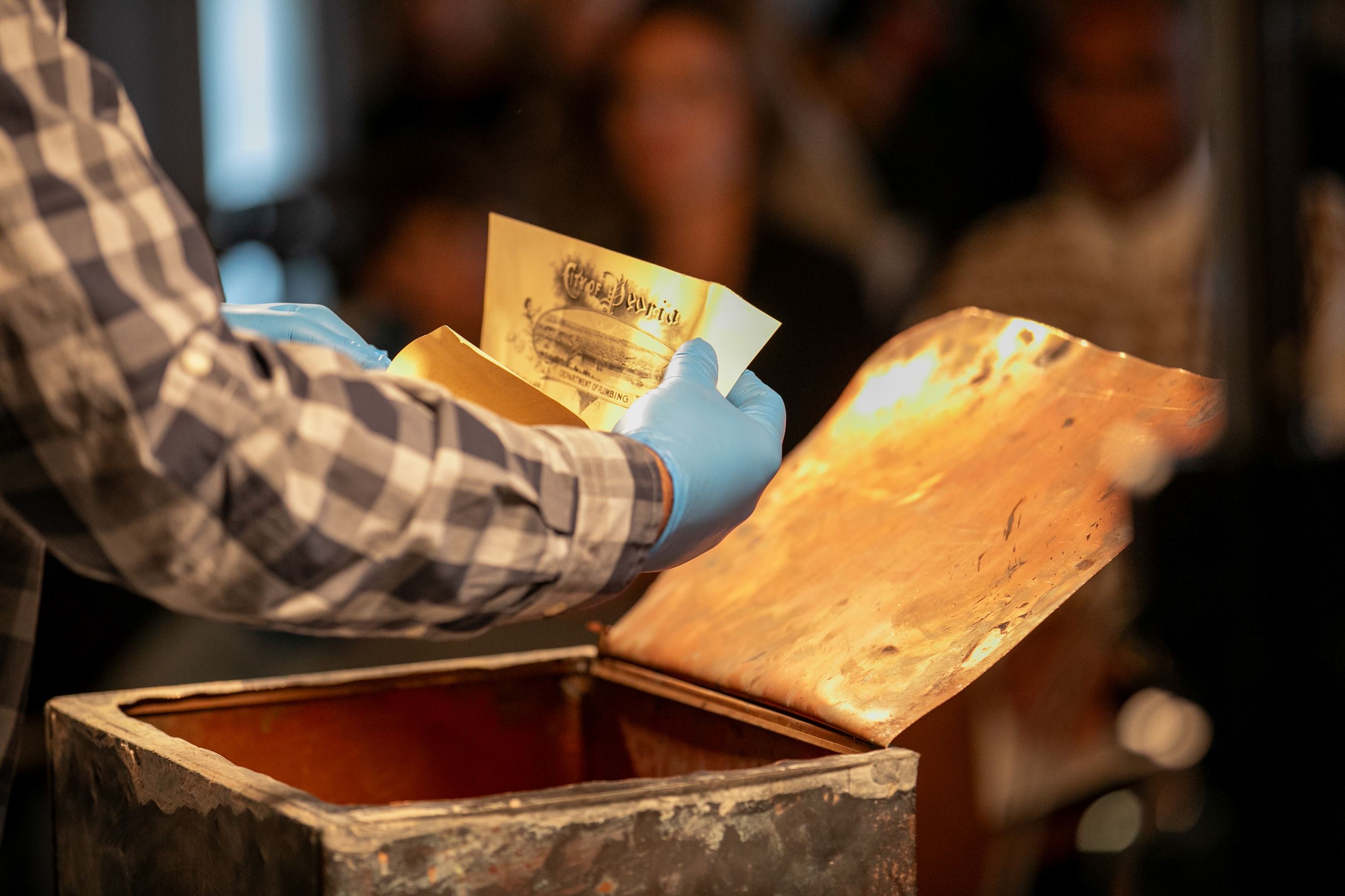 Gloved hands hold a document over a time capsule that reads "City of Peoria" at the top.